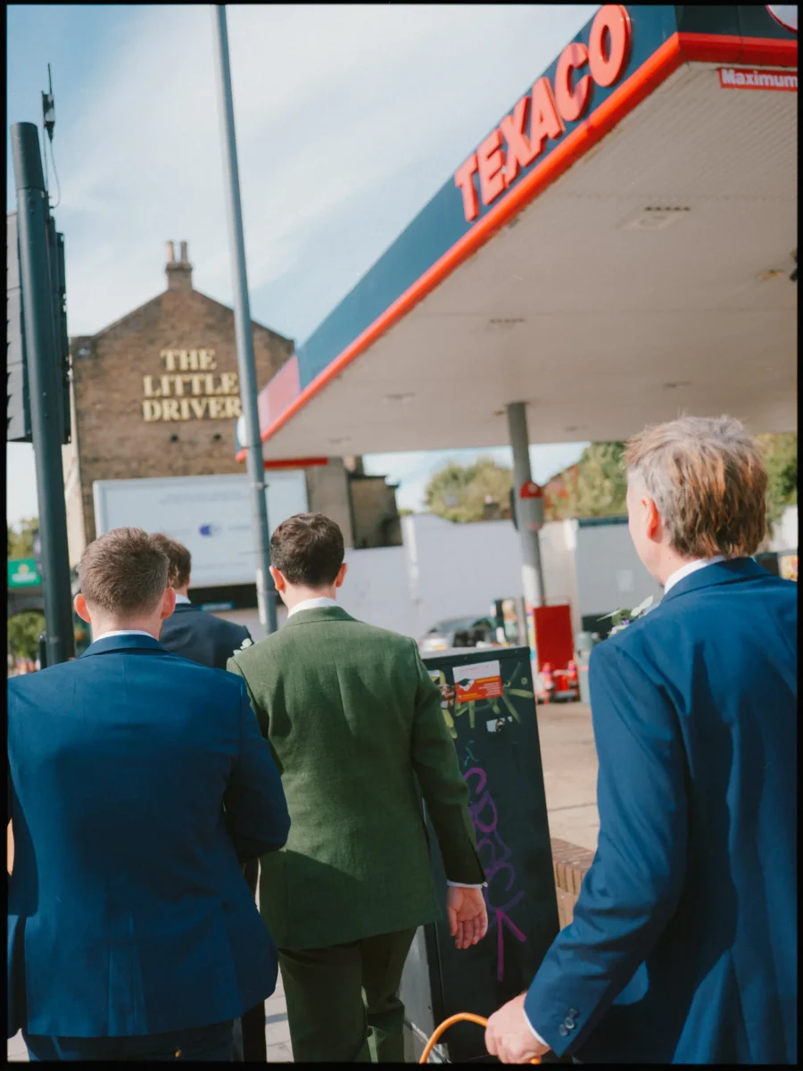 Groomsmen in blue and green suits walk past a Texaco garage towards The Little Driver pub on the way to Bow Church