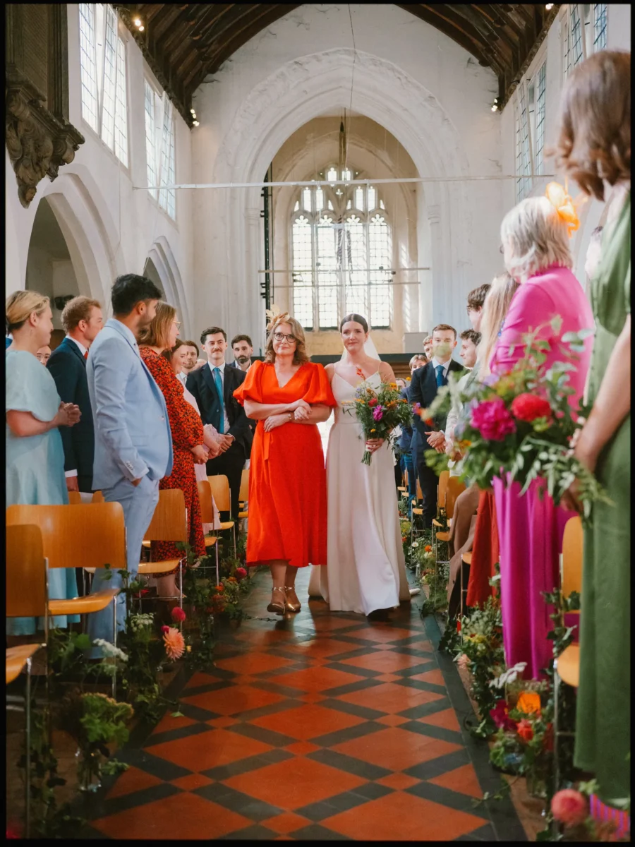 Bride in a white dress walks down the aisle at Bow Church on the arm of a woman in an orange dress, guests smiling
