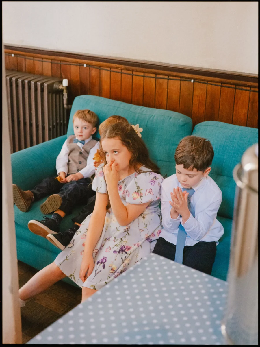 Children in wedding outfits on a teal sofa, a girl in a floral dress hand to mouth, a boy with hands together
