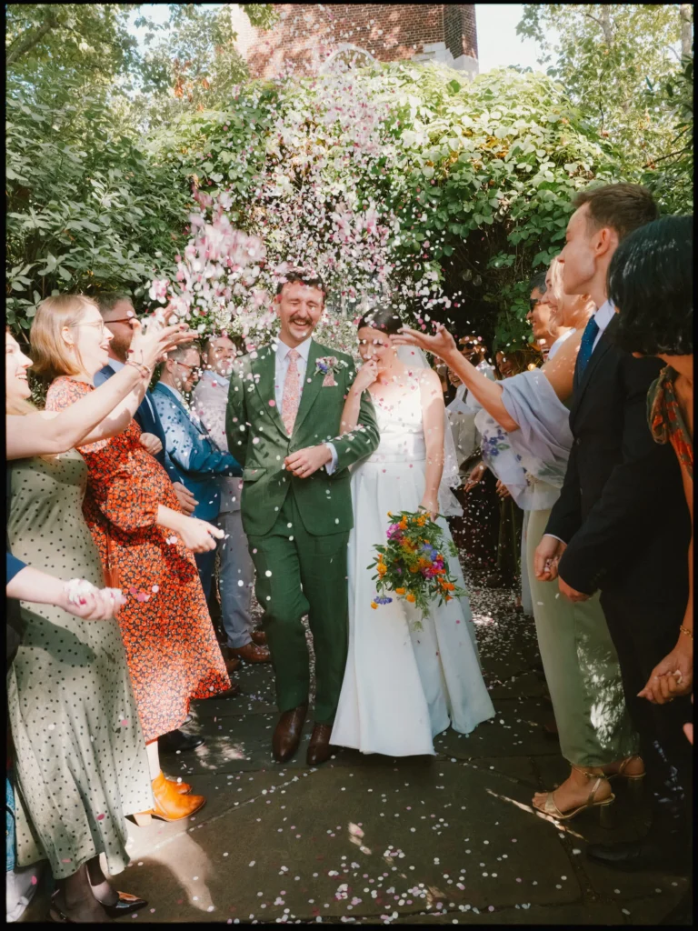 Laughing groom in a green suit and bride with wildflower bouquet walk through guests showering them in confetti at Bow Church