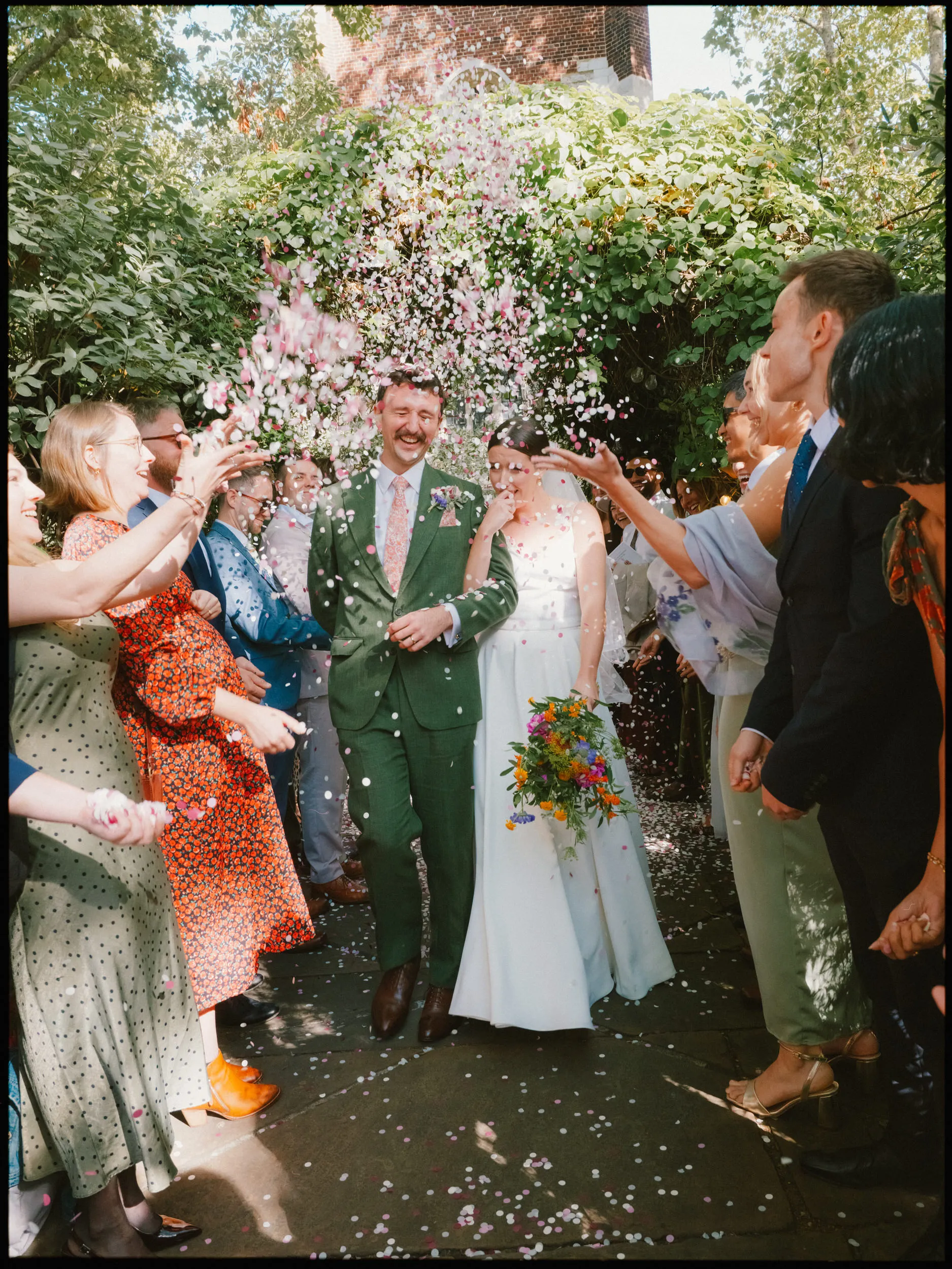 Laughing groom in a green suit and bride with wildflower bouquet walk through guests showering them in confetti at Bow Church