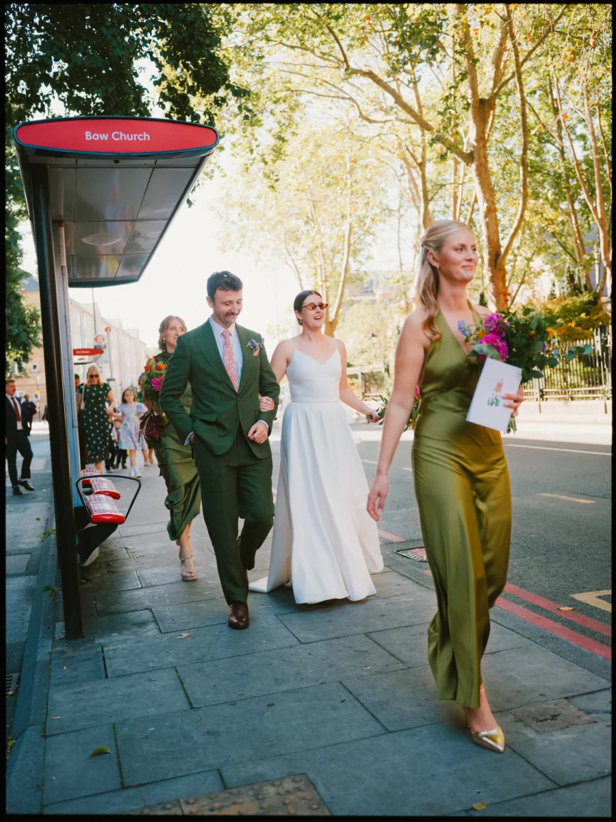 Bride in sunglasses and groom in green suit stroll arm in arm past the Bow Church bus stop with their bridesmaids