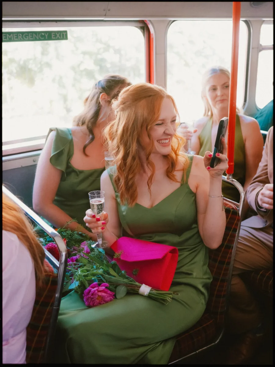 Bridesmaid in an olive green dress laughs on the bus, holding champagne and a phone, bouquet and pink clutch in her lap