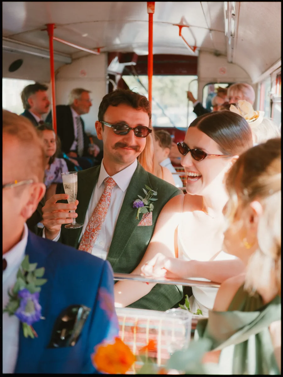 Moustached groom in sunglasses raises a champagne glass as the laughing bride leans in, guests packed on the bus
