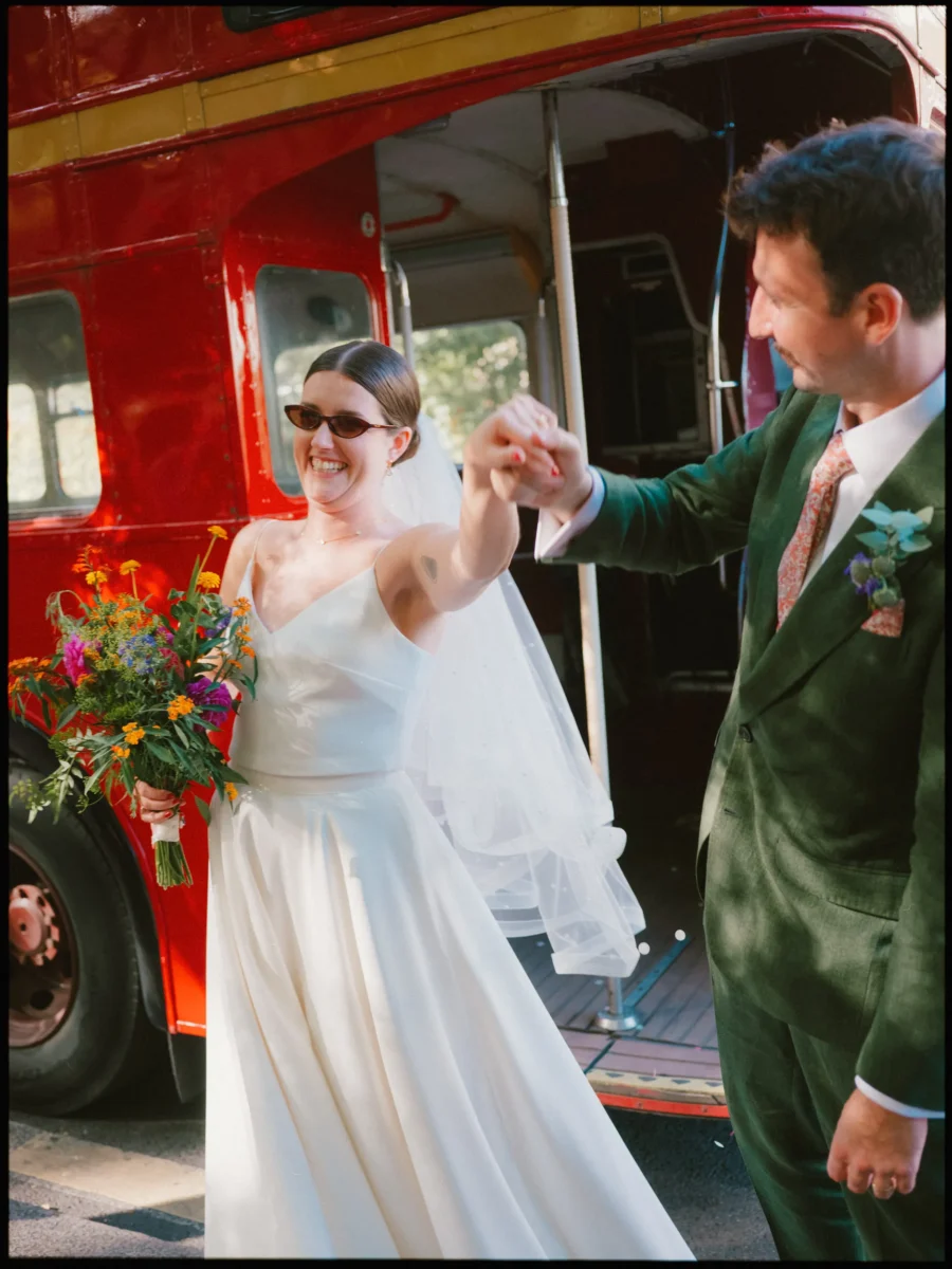 Smiling bride in sunglasses holds a wild flower bouquet and takes her groom's hand as they step off a red London bus