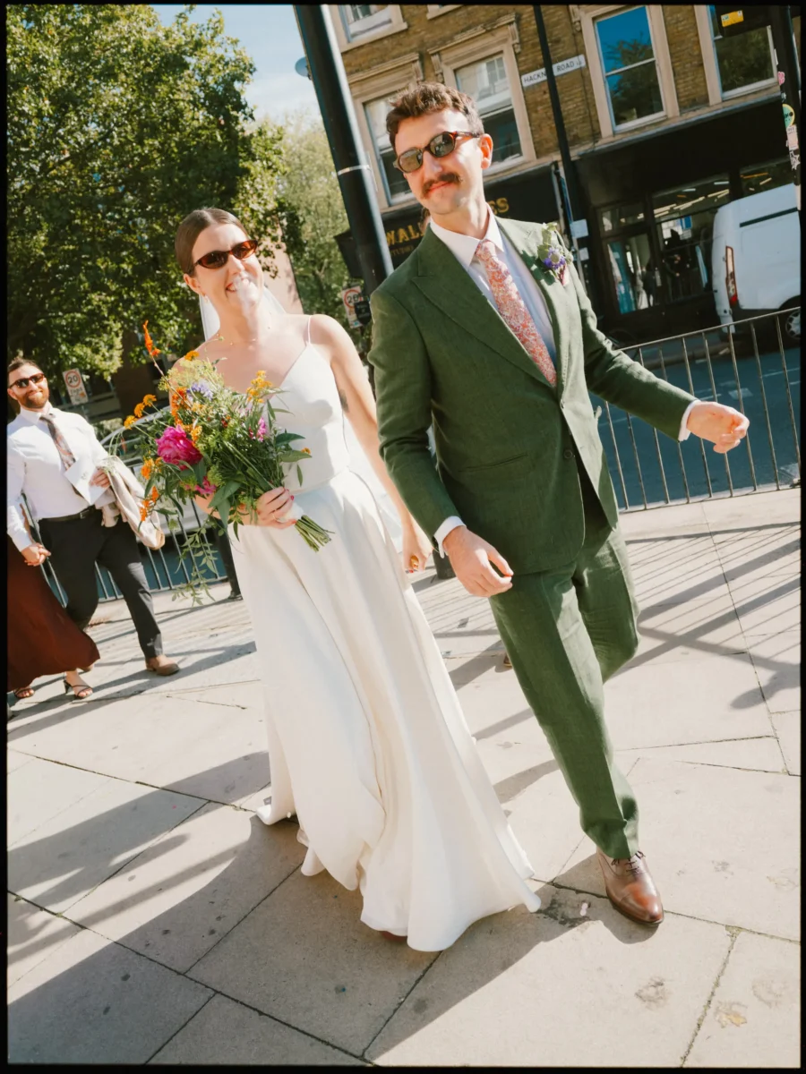 Bride and groom in matching sunglasses stride along a sunlit Hackney street, her bright bouquet in hand, guests following