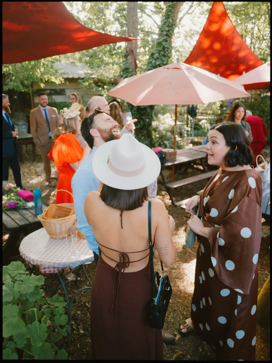 Guests chat and laugh under red sails and a pink parasol in the dappled sunlight of Hackney City Farm's garden