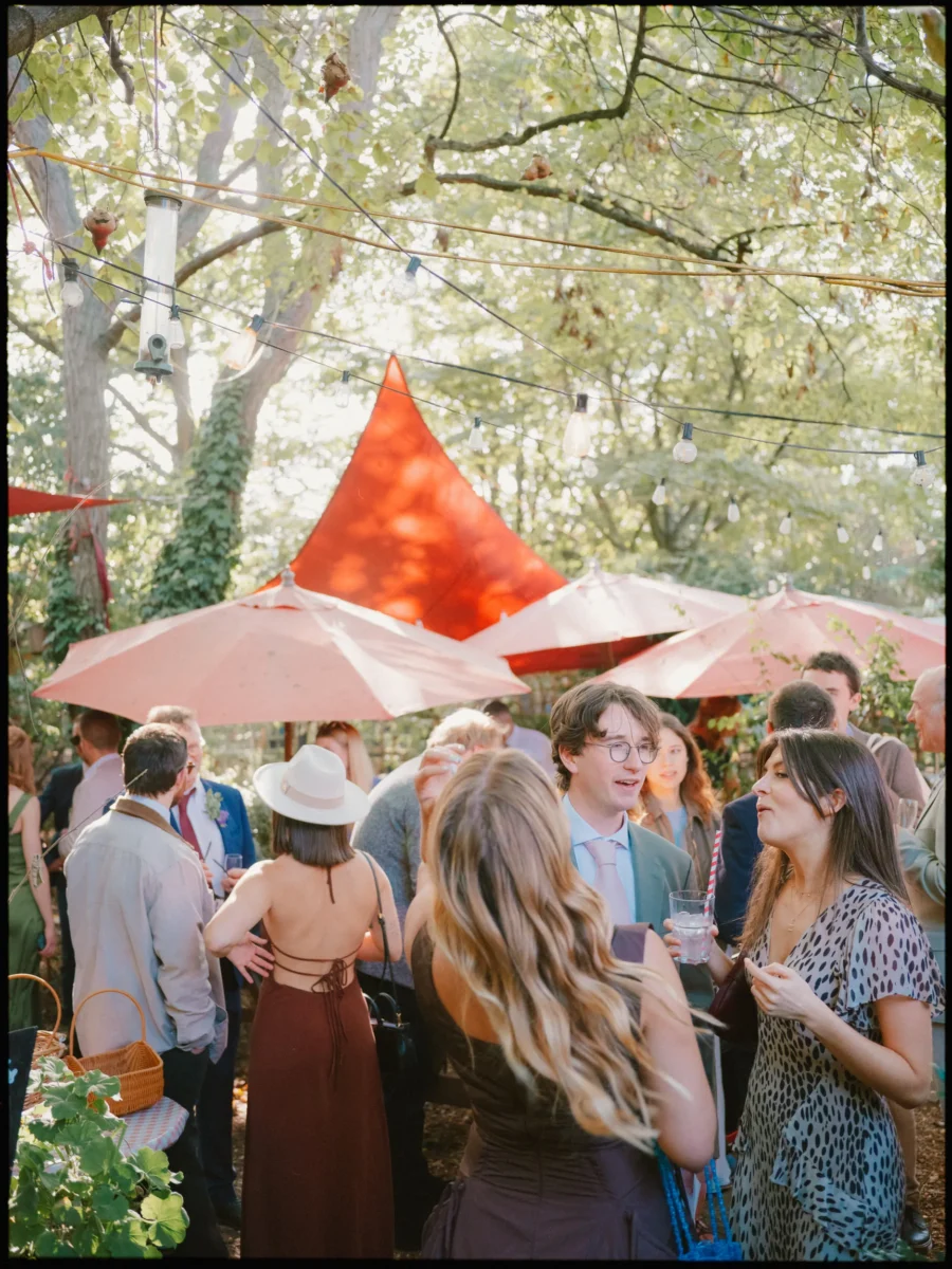 Wedding guests mingling with drinks beneath festoon lights and peach parasols, a woman in a straw hat seen from behind