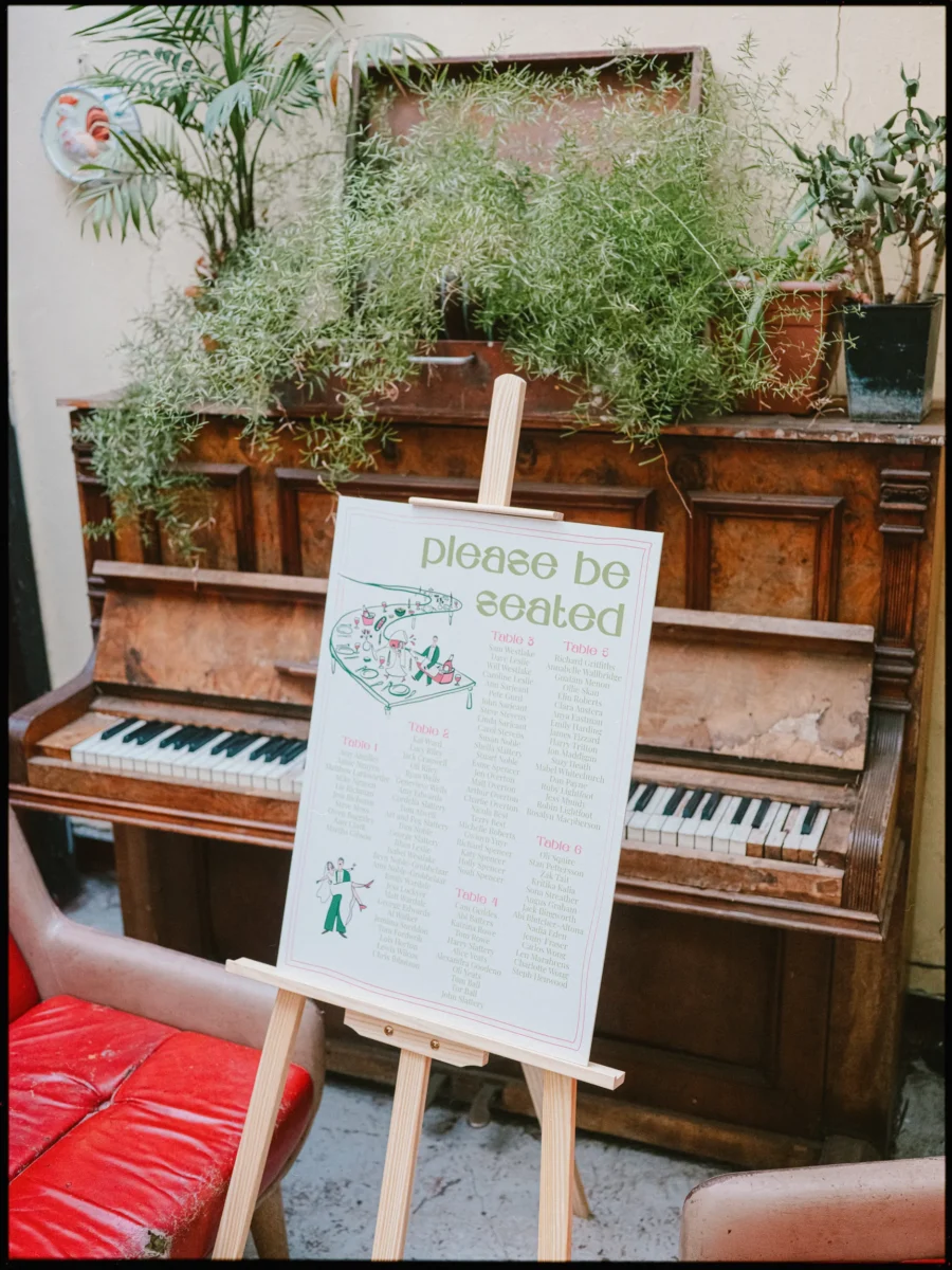 Illustrated 'Please be seated' table plan on a wooden easel beside an old upright piano at Hackney City Farm