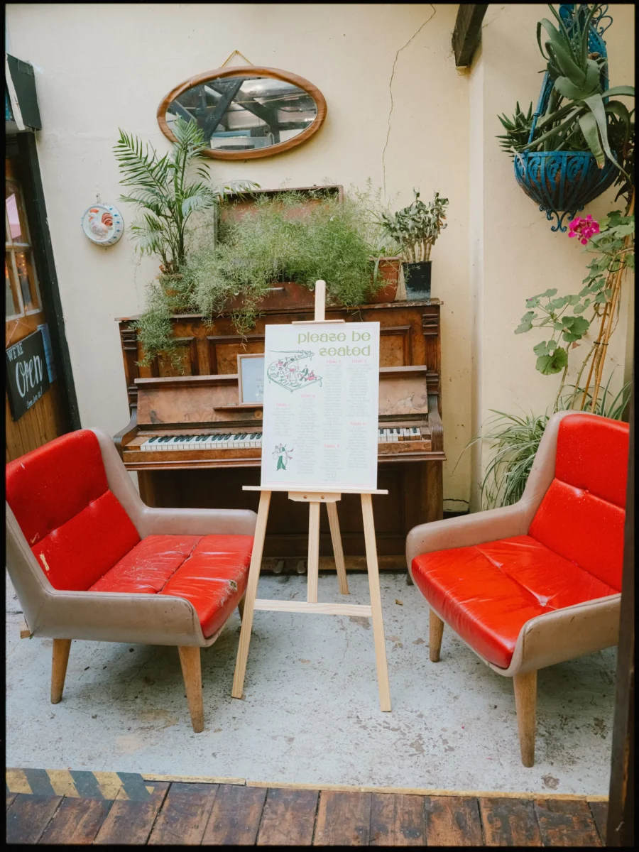 Two worn red leather armchairs either side of the seating plan easel, with an old piano and trailing plants at Frizzante