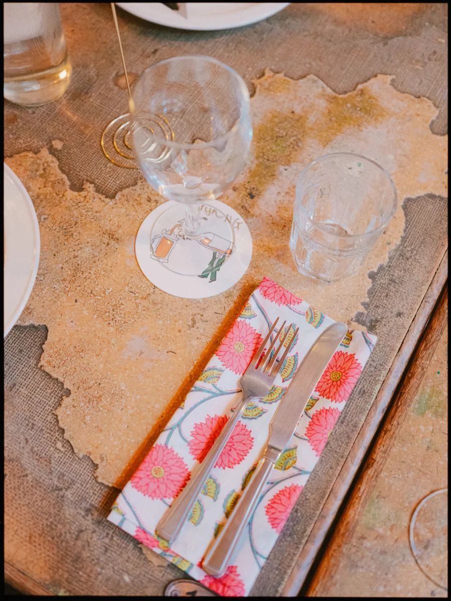 Pink floral napkin with a fork and knife, a wine glass on a pub-style coaster and tumbler on a weathered table at Frizzante