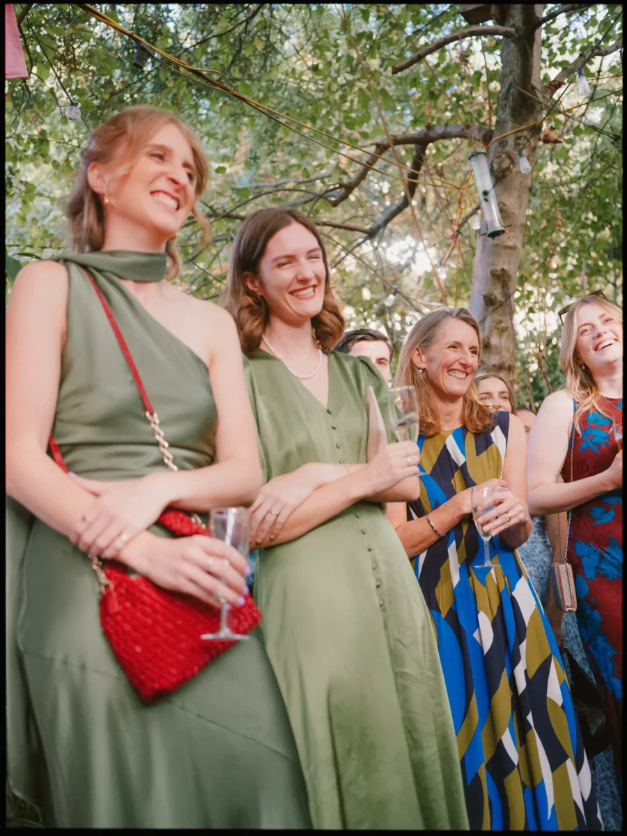 Four women guests laugh together under the trees, glasses in hand, listening to the speeches at Hackney City Farm