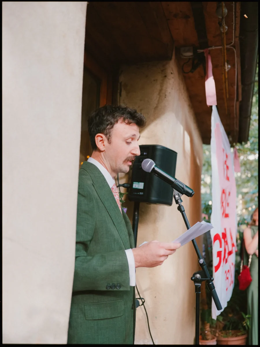 Moustached man in a green linen suit reads his speech from a sheet of paper at a microphone outside, banner behind