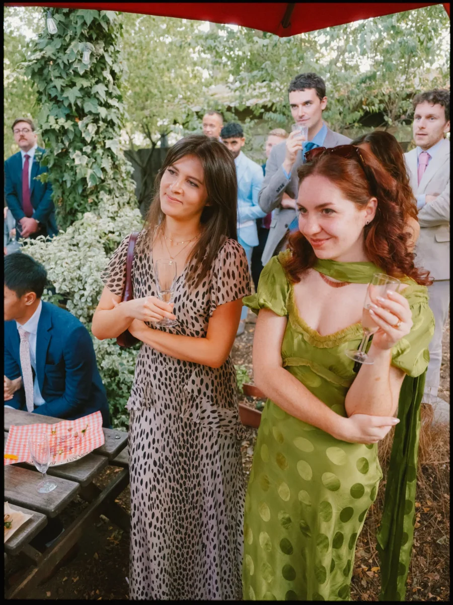 Two women guests listen to the speeches outdoors, glasses in hand, one in a leopard print dress, one in green polka dots