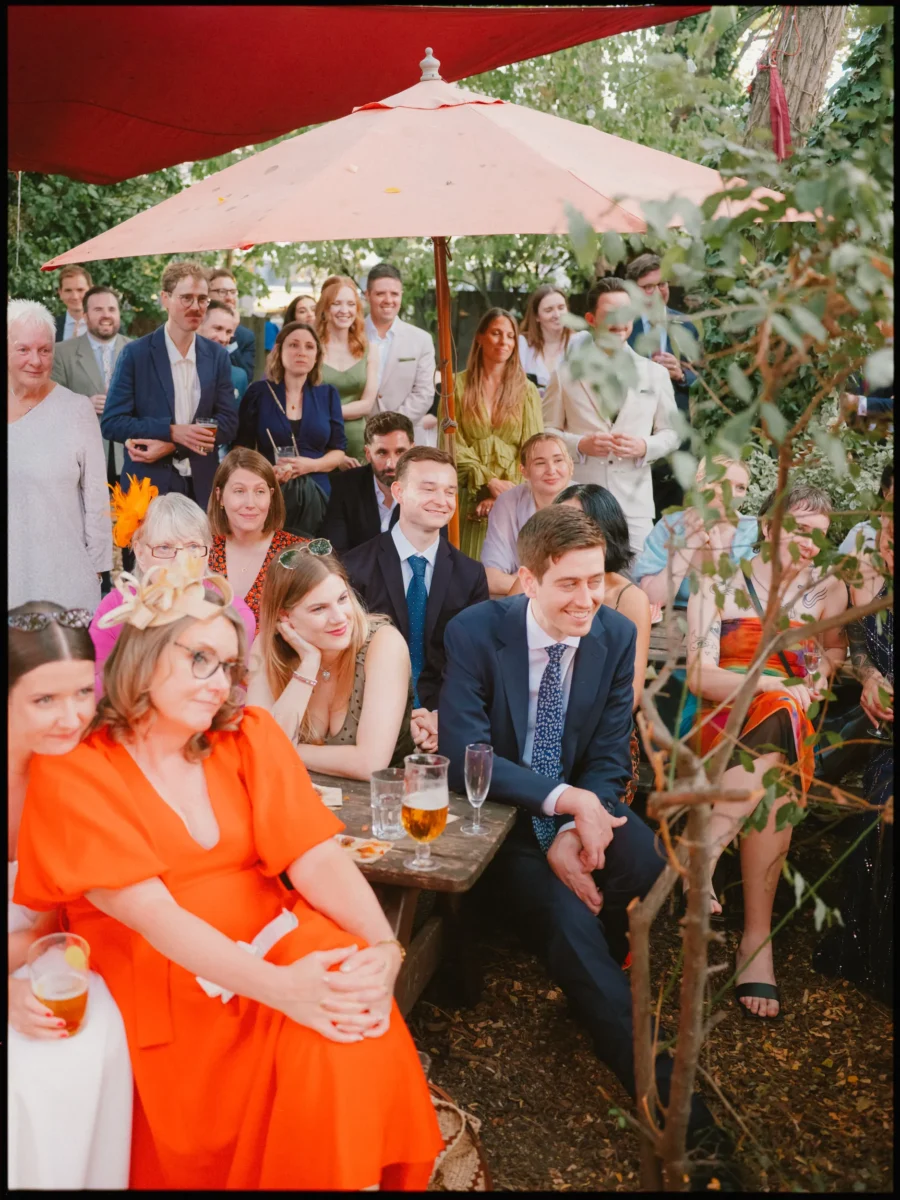 Guests crowd together under parasols at Hackney City Farm, smiling and listening to outdoor wedding speeches