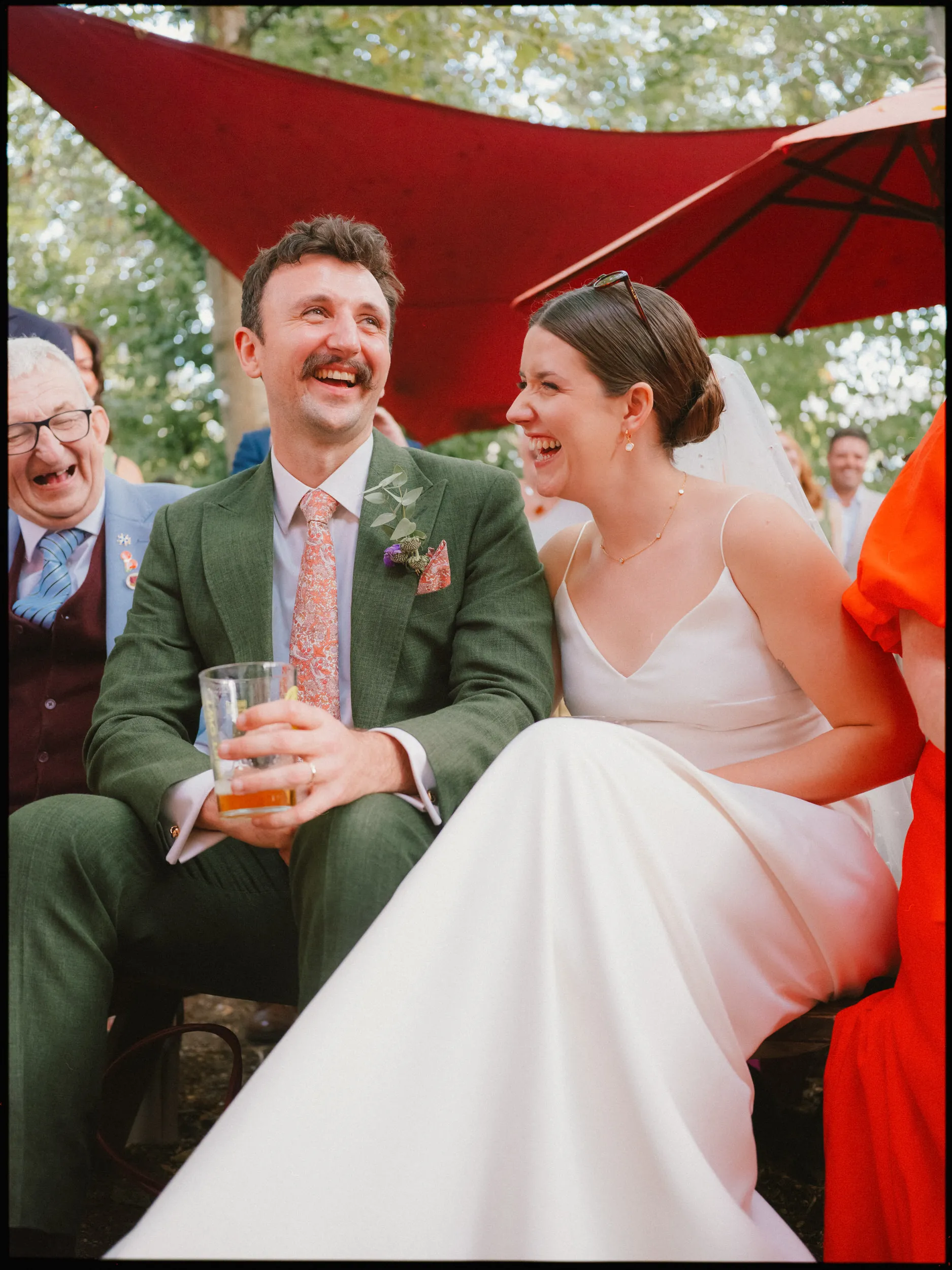 Bride and groom laugh together on a bench during the outdoor speeches, he holding a pint, red parasols above them