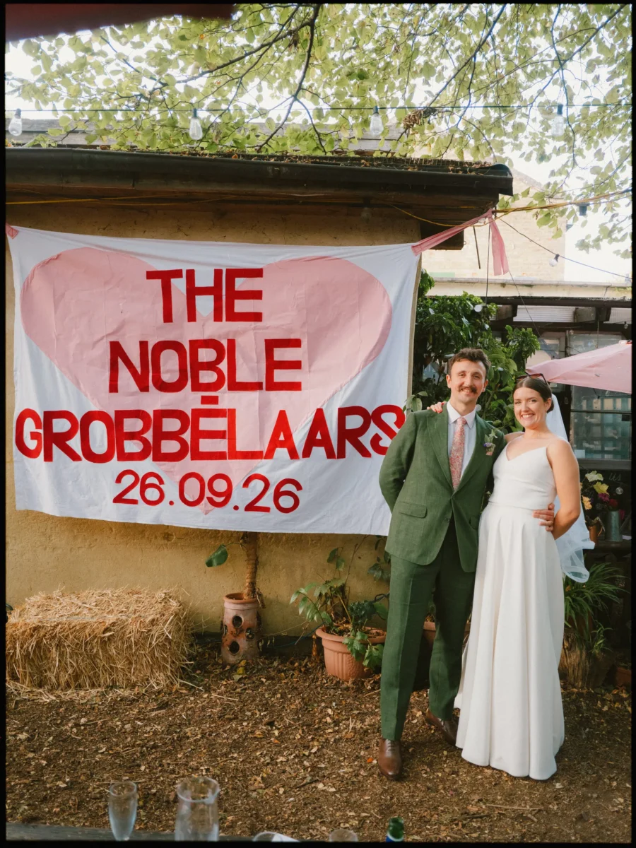 Bride in a white dress and groom in a green suit stand arm in arm beside a hand-painted heart banner at Hackney City Farm
