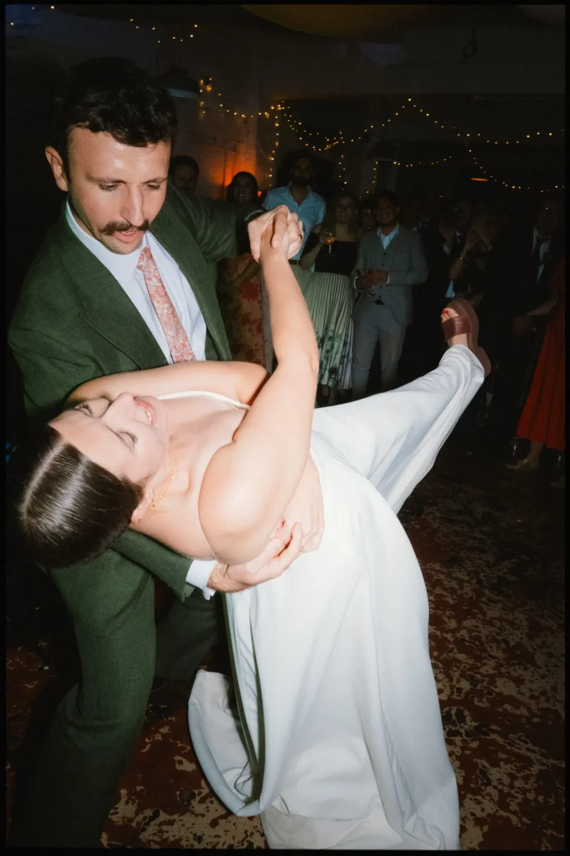 Groom in a green suit dips the bride low, her foot kicked up, as guests watch from the Frizzante dance floor