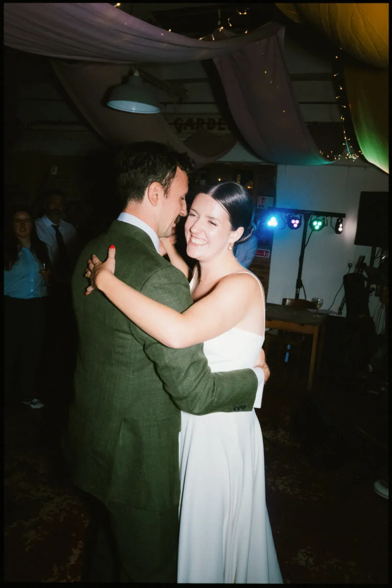 Bride laughing with her eyes shut as she dances close with the groom in his green suit, stage lights glowing behind