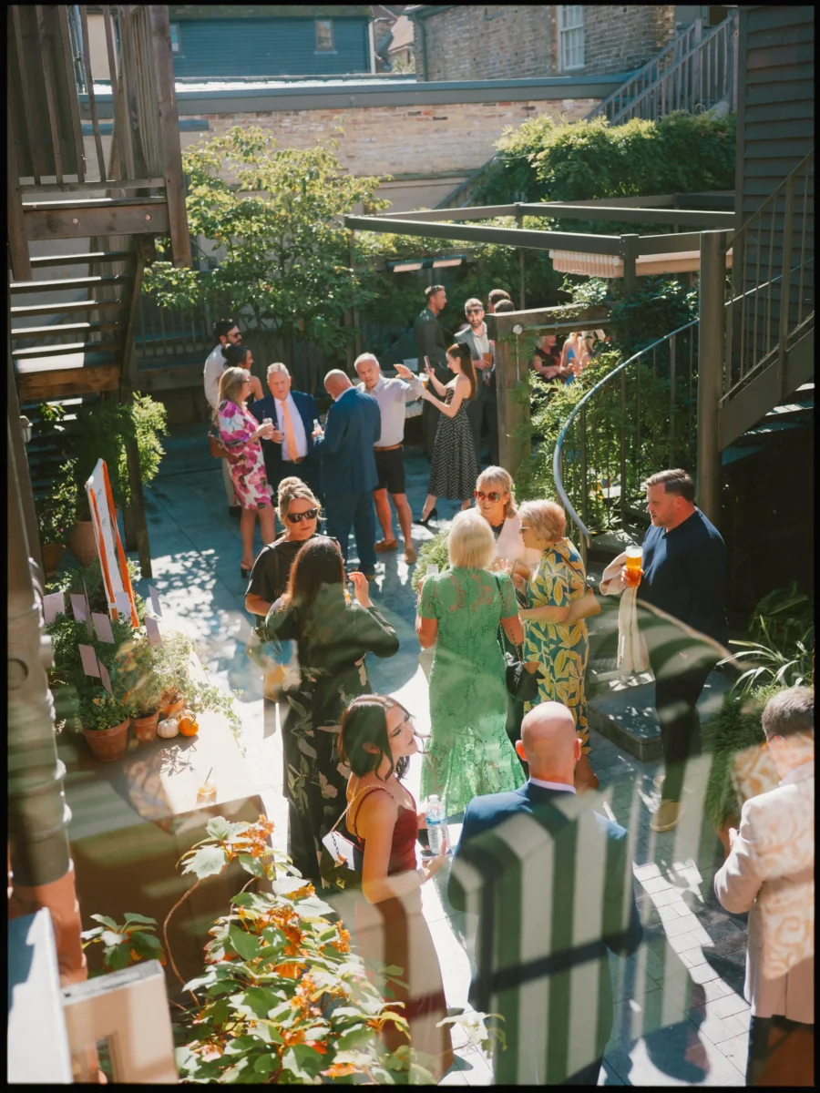Guests in summer dresses and suits chat with drinks on the sun-dappled courtyard at The George, seen from a stairway