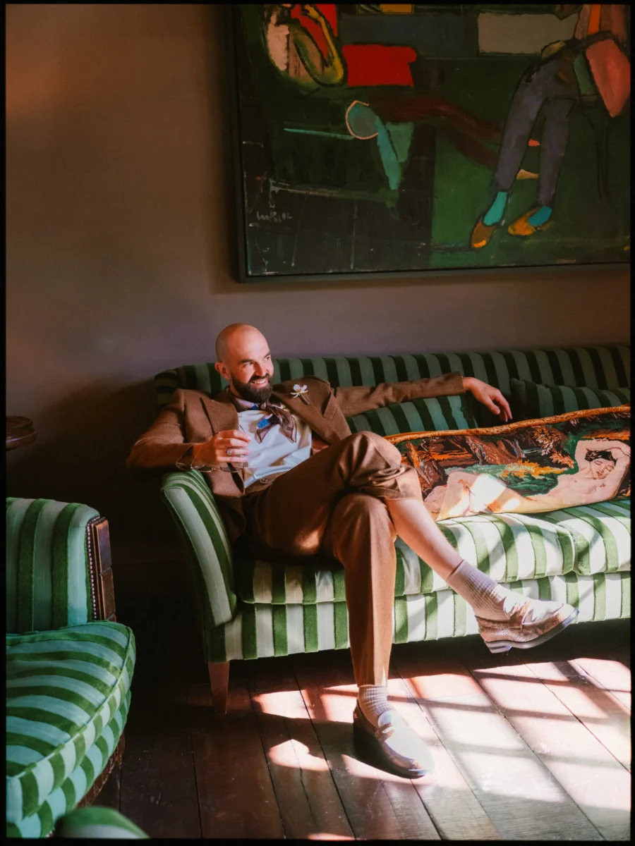 Ross in a brown suit and bow tie relaxes on a green striped sofa in the sunlit green room at The George, smiling