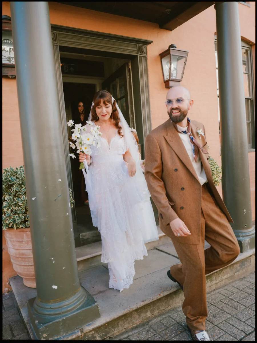 Bride with a white flower bouquet and groom in a brown suit step smiling out of The George's columned doorway