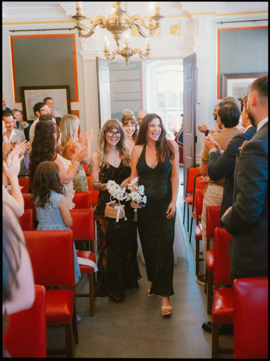 Two women in dark dresses carry white daisies down the aisle, laughing as standing guests applaud at Rye Town Hall