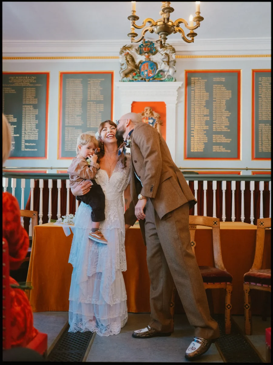Laughing bride holds a little boy as her groom in a brown suit kisses her cheek in the Rye Town Hall ceremony room