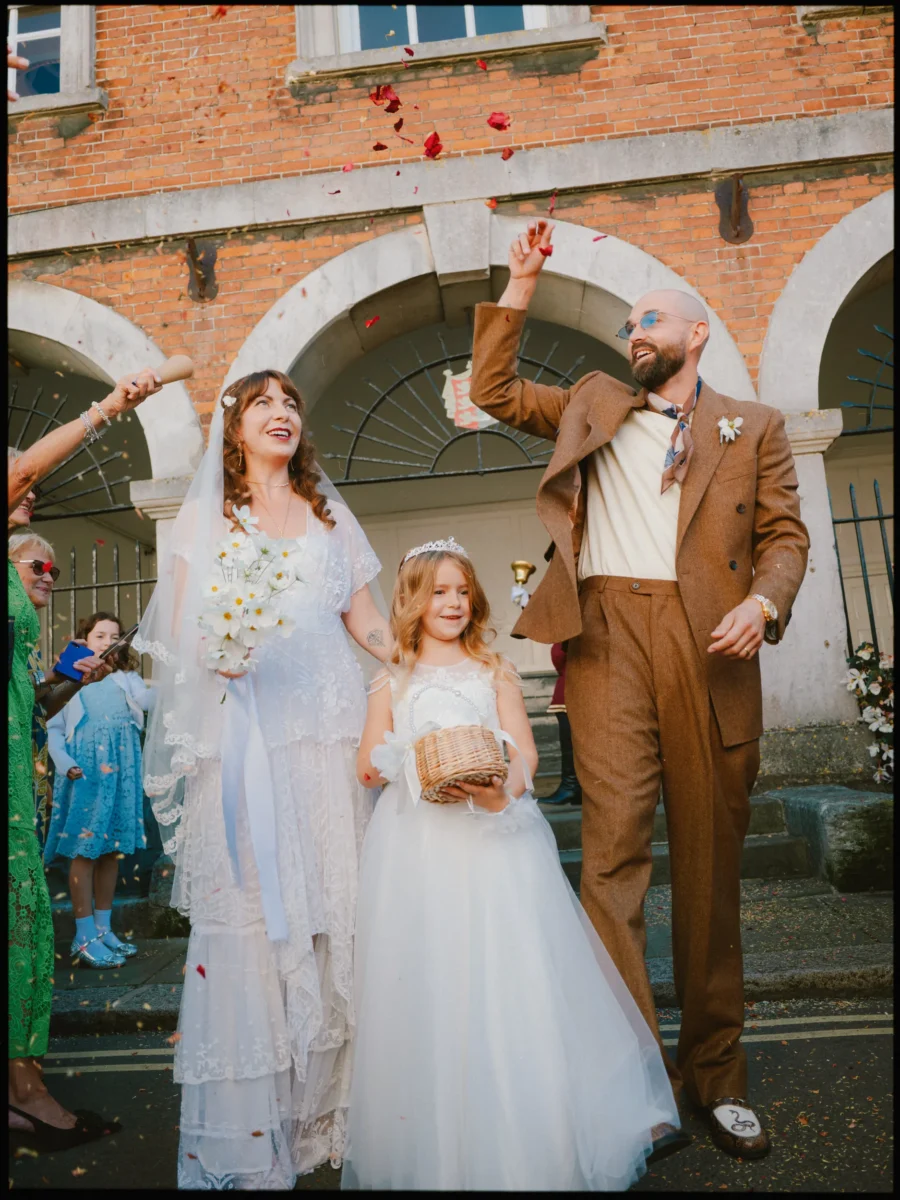 Bride, groom and flower girl laugh as guests throw red petal confetti outside Rye Town Hall's arched brick entrance
