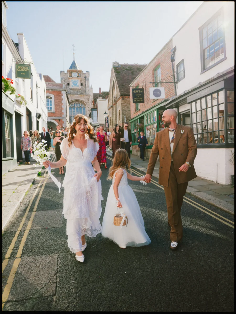 Smiling bride in a lace dress and groom in a brown suit walk hand in hand with a flower girl down a Rye street
