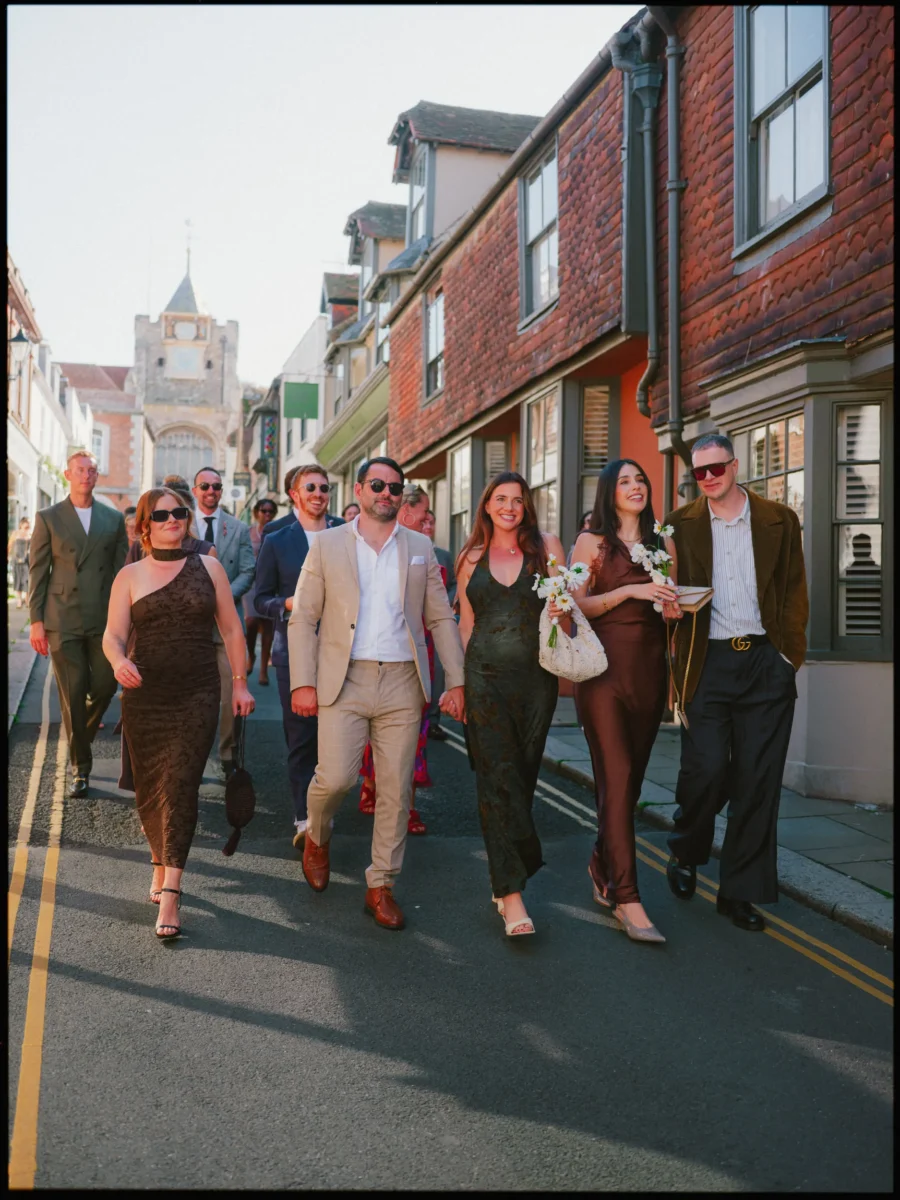 Smiling guests in sunglasses and rich autumn colours stroll down a sunlit Rye street, carrying white daisies