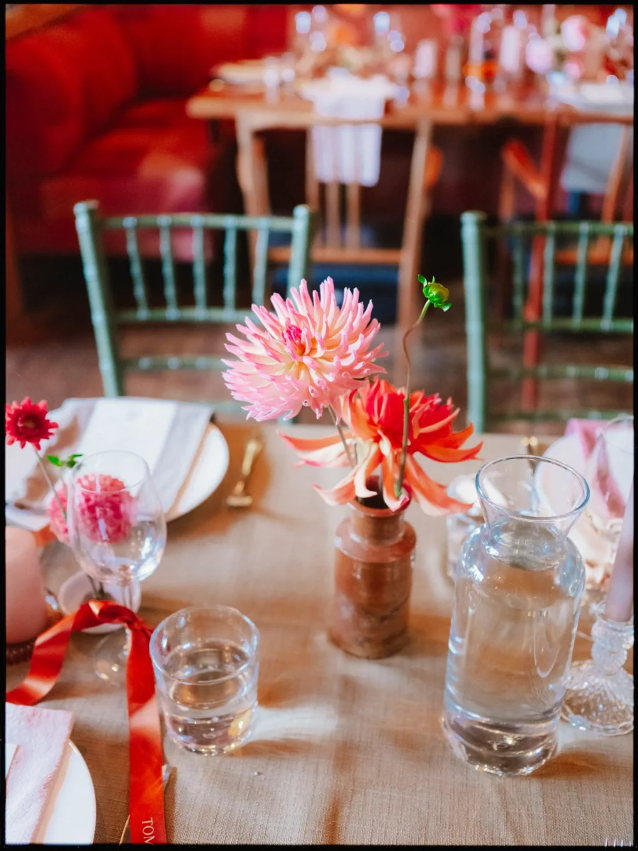 Pink and coral dahlias in a small terracotta vase on a hessian table runner, with a water carafe and orange ribbon