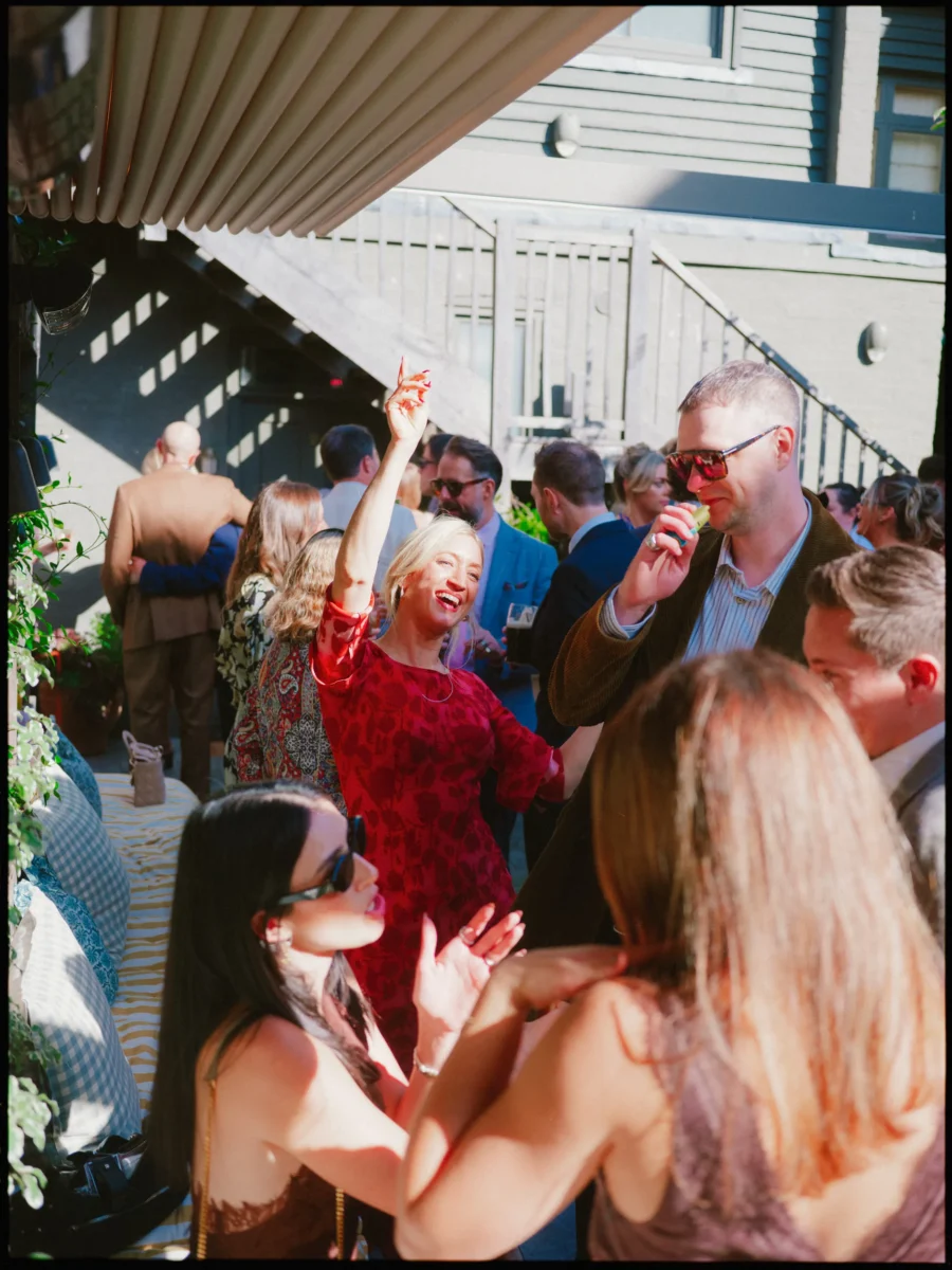 Guest in a red patterned dress laughs and dances, hand in the air, as friends in sunglasses mingle in afternoon sun