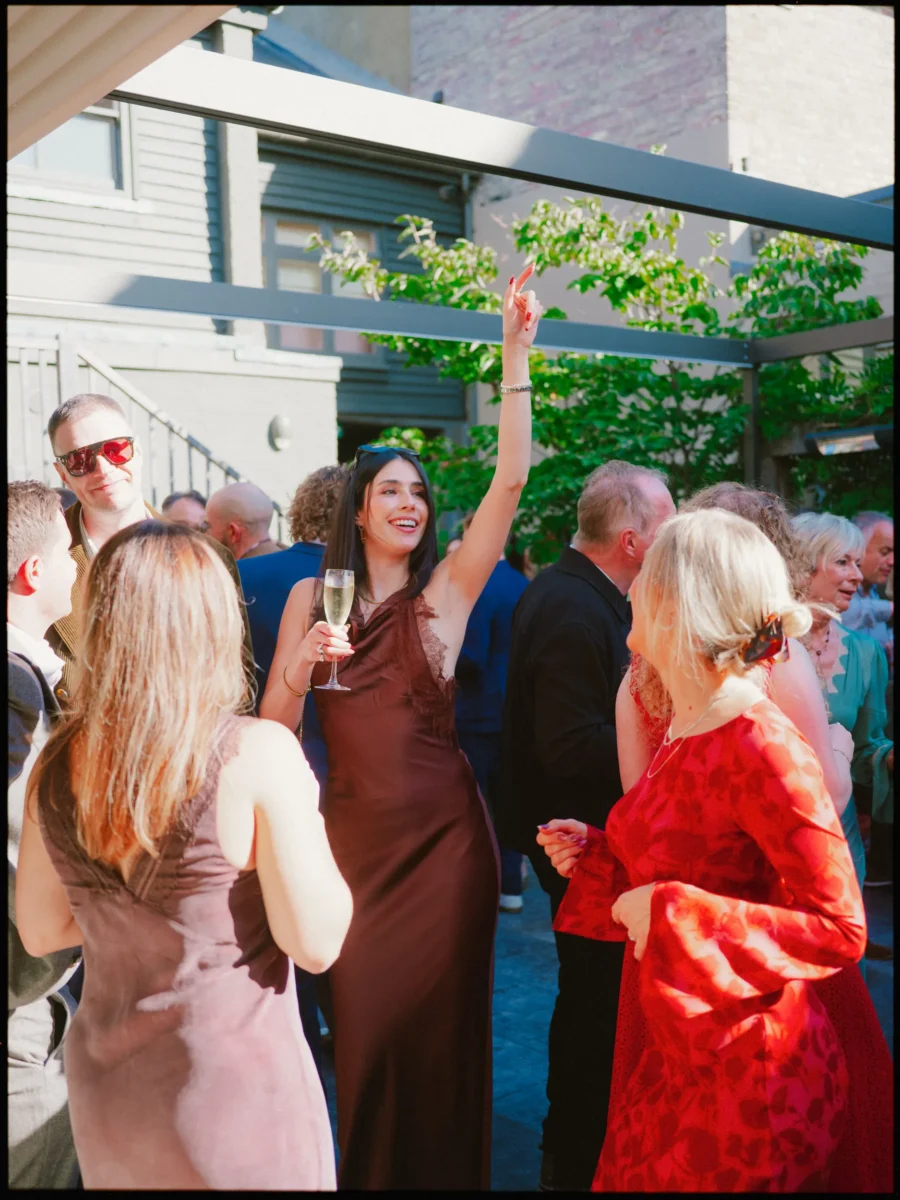 Woman in a burgundy satin dress laughs with one arm raised and a champagne flute in hand, among guests on a sunny terrace