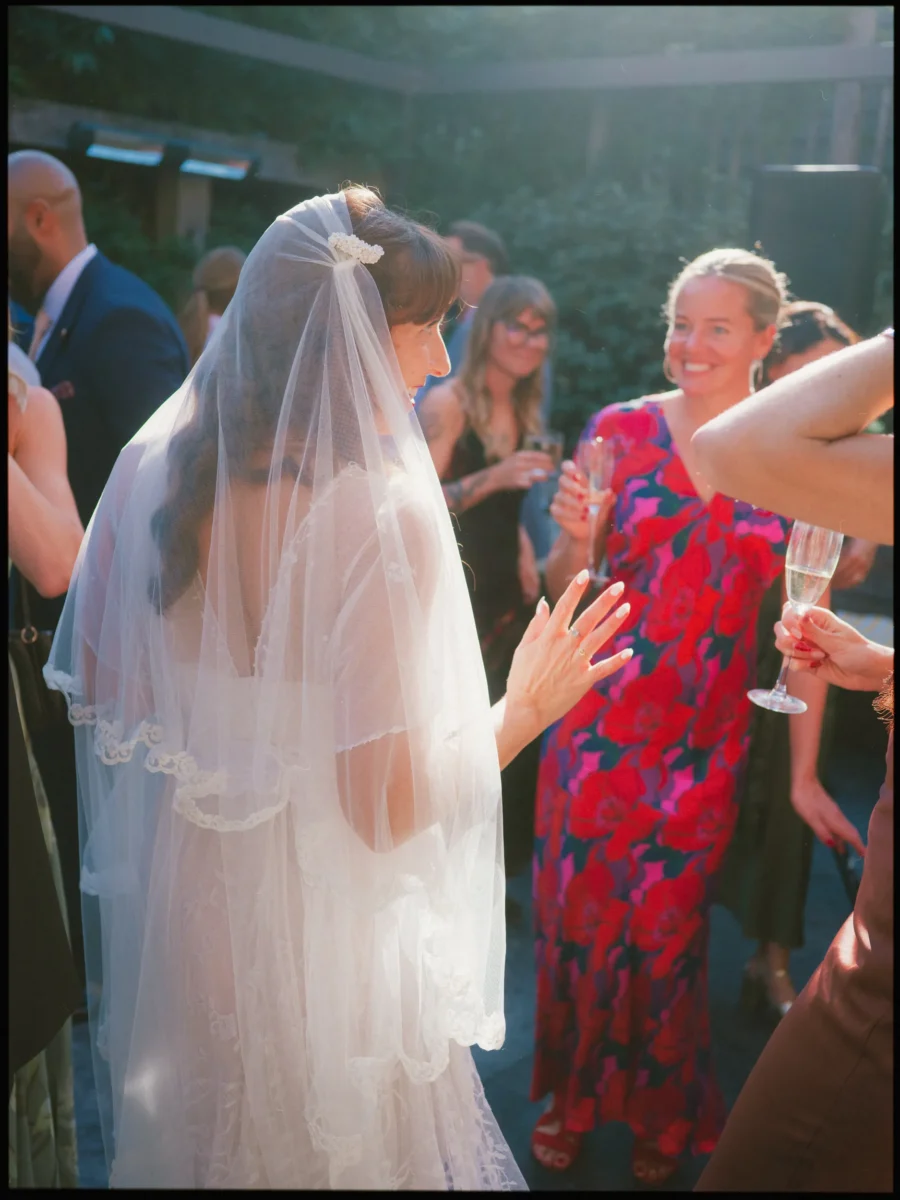 Bride in lace-trimmed veil shows her ring to a smiling guest holding champagne in the sunny courtyard