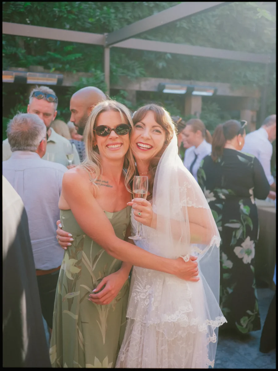 Bride in a lace veil hugs a laughing guest in sunglasses and a green dress, champagne in hand, at The George