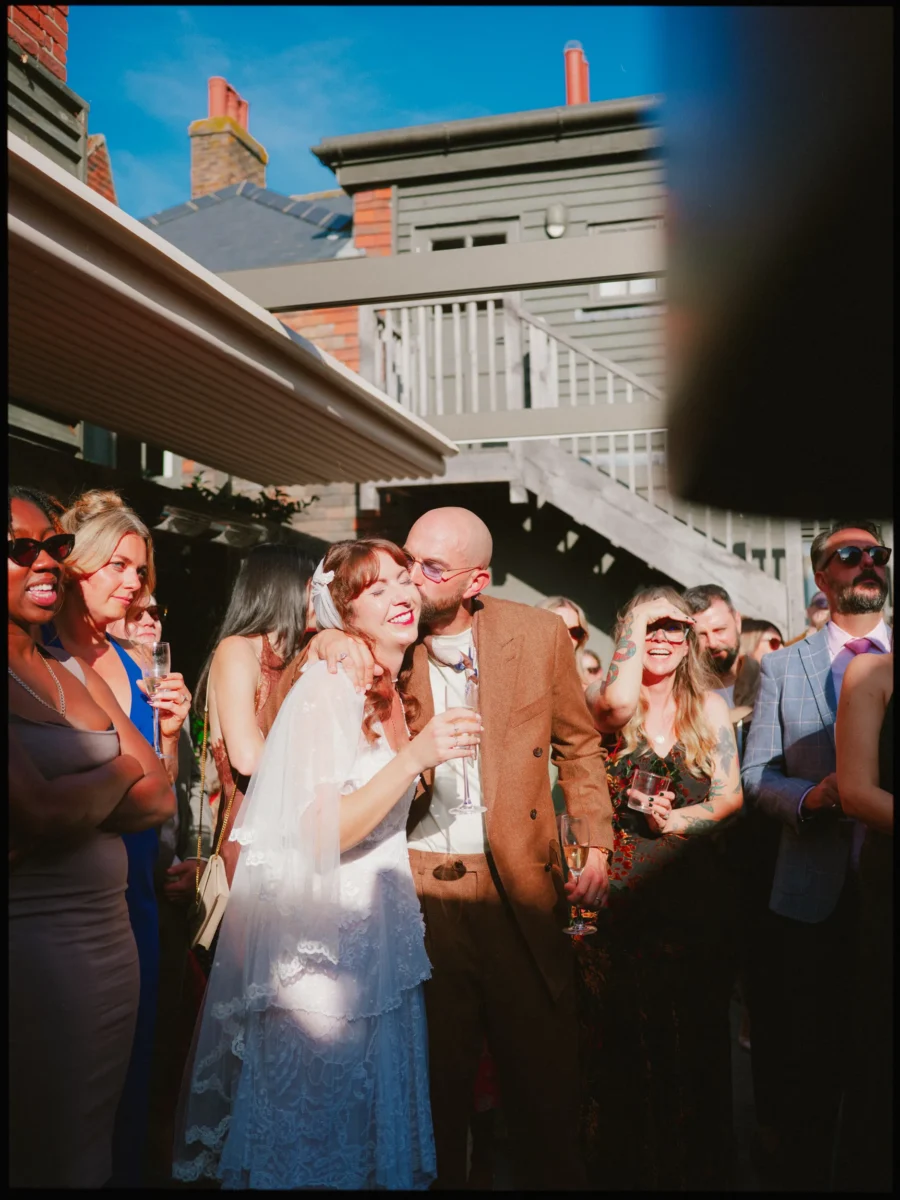 Bride laughing as her groom kisses her cheek, surrounded by guests with drinks in the sunny courtyard at The George