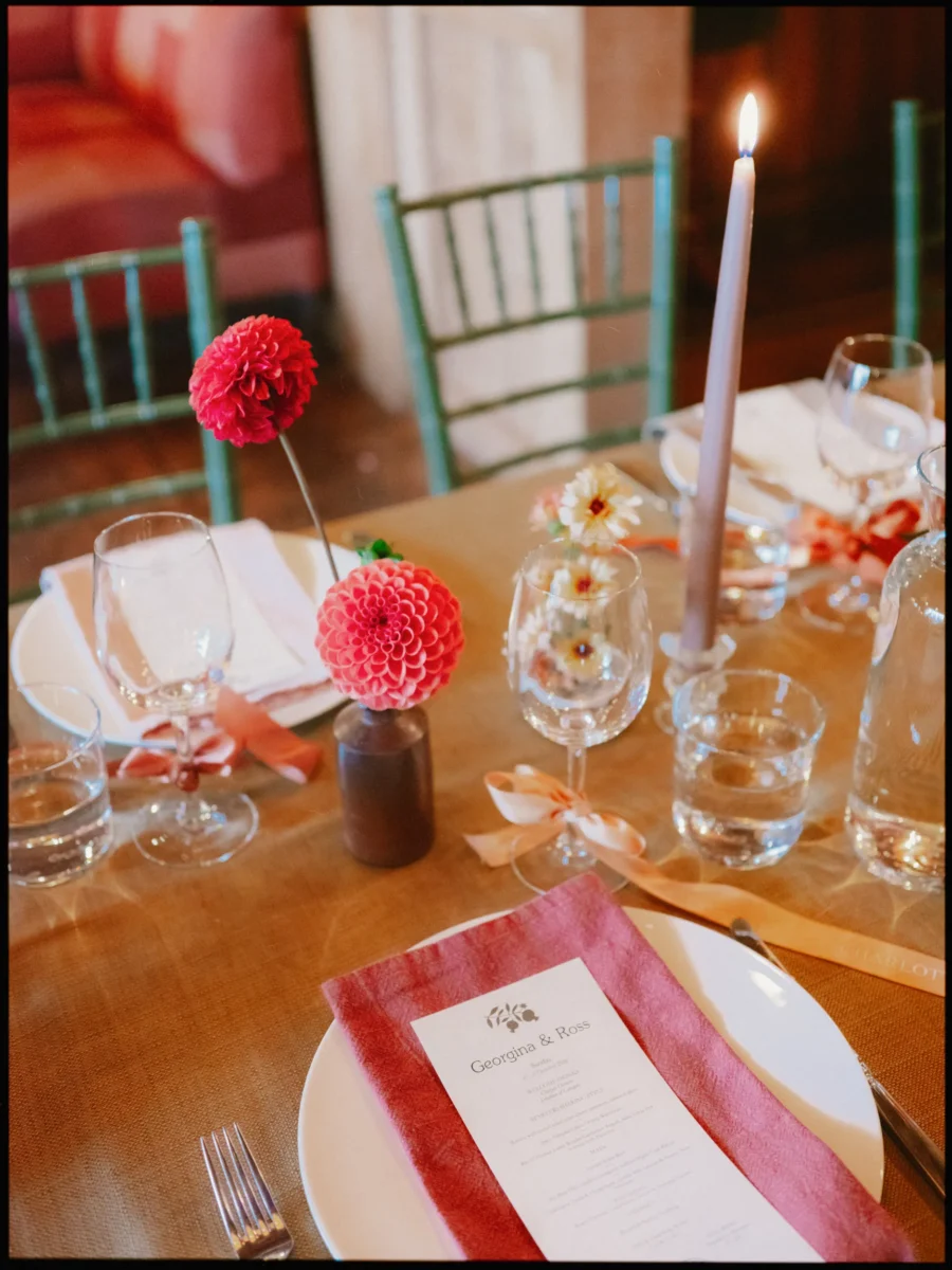 Lit taper candle, glasses and a red dahlia in a small vase beside a Georgina & Ross menu on a rose napkin