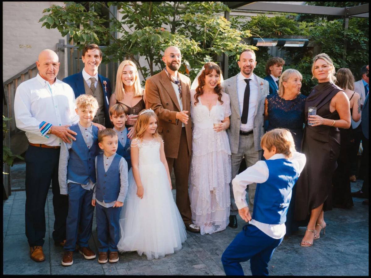Bride and groom smile with family in a sunlit courtyard at The George in Rye as a boy in a blue waistcoat dances