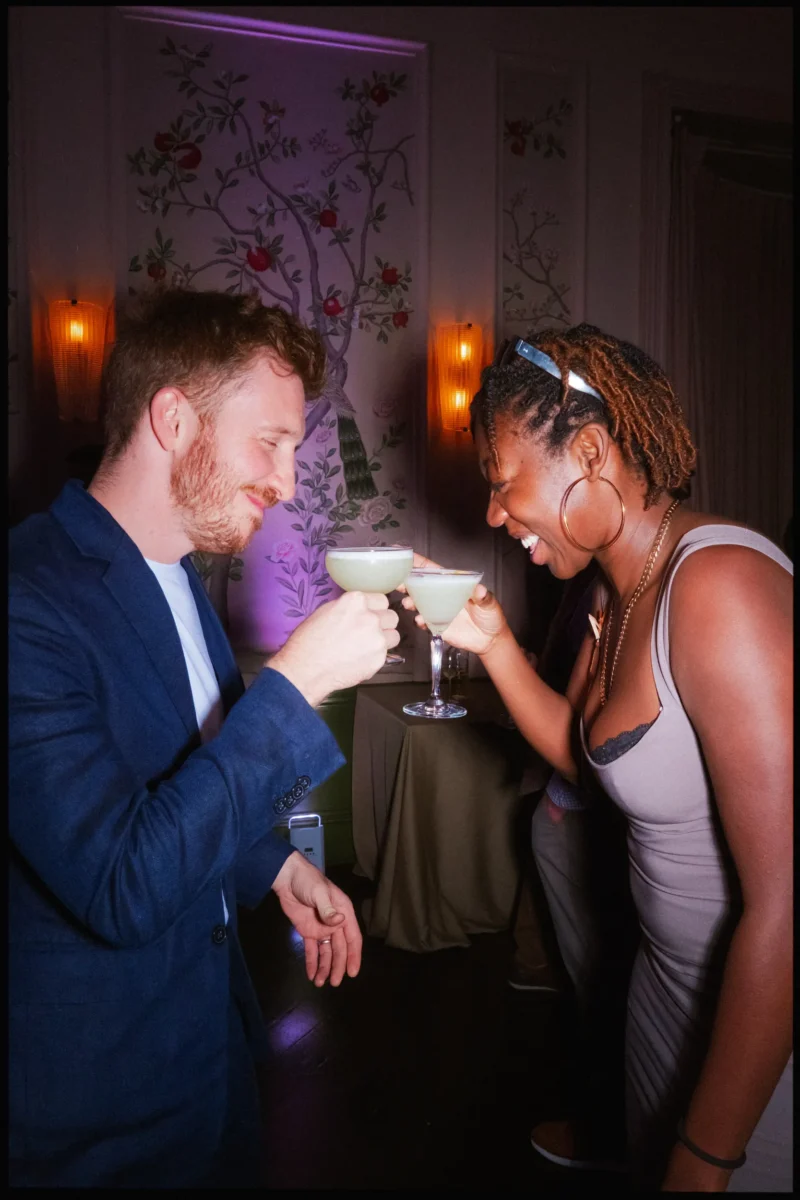 Laughing guests clink pale green cocktails beside glowing wall lights and floral painted panels at The George