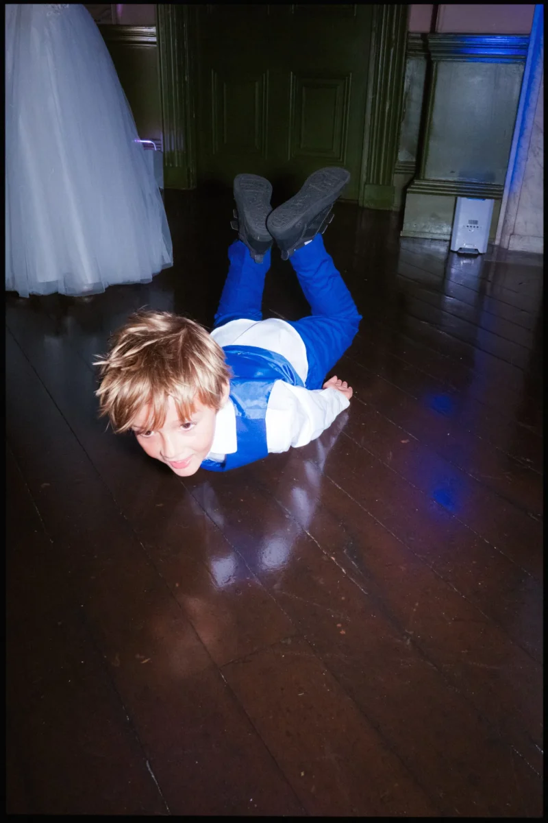 Little boy in a bright blue waistcoat slides across the polished wooden floor on his tummy, grinning, feet in the air