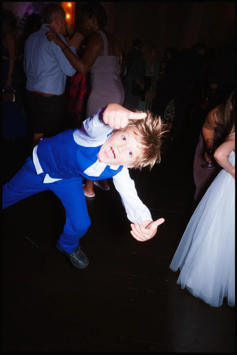 Boy in a bright blue waistcoat leans sideways with thumbs pointing at his head, tongue out, on the dark dancefloor