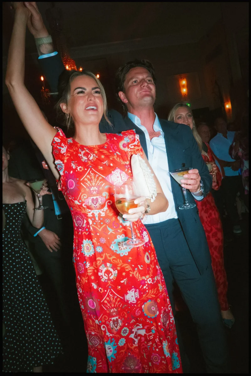 Guest in a red floral dress punches the air with a cocktail, dancing beside a man in navy in The George Ballroom