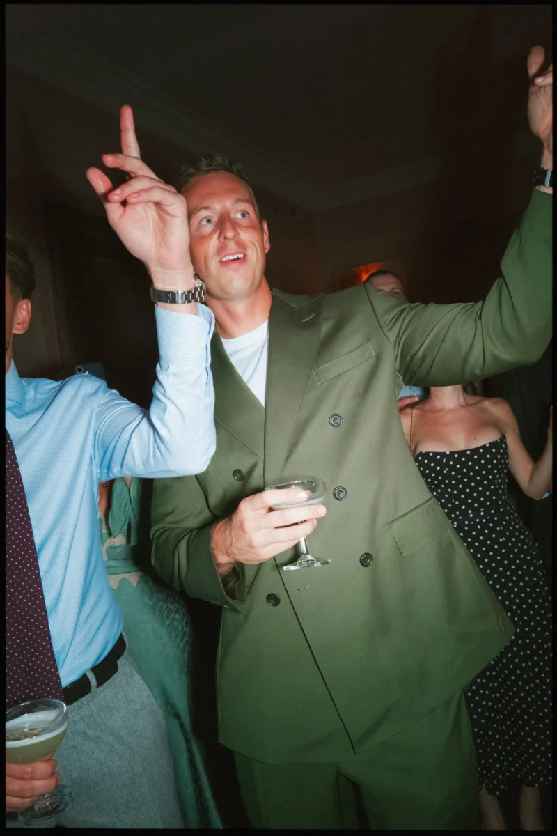 Guest in a green double-breasted suit holds a coupe glass and points upwards, mid-song on the dancefloor at The George