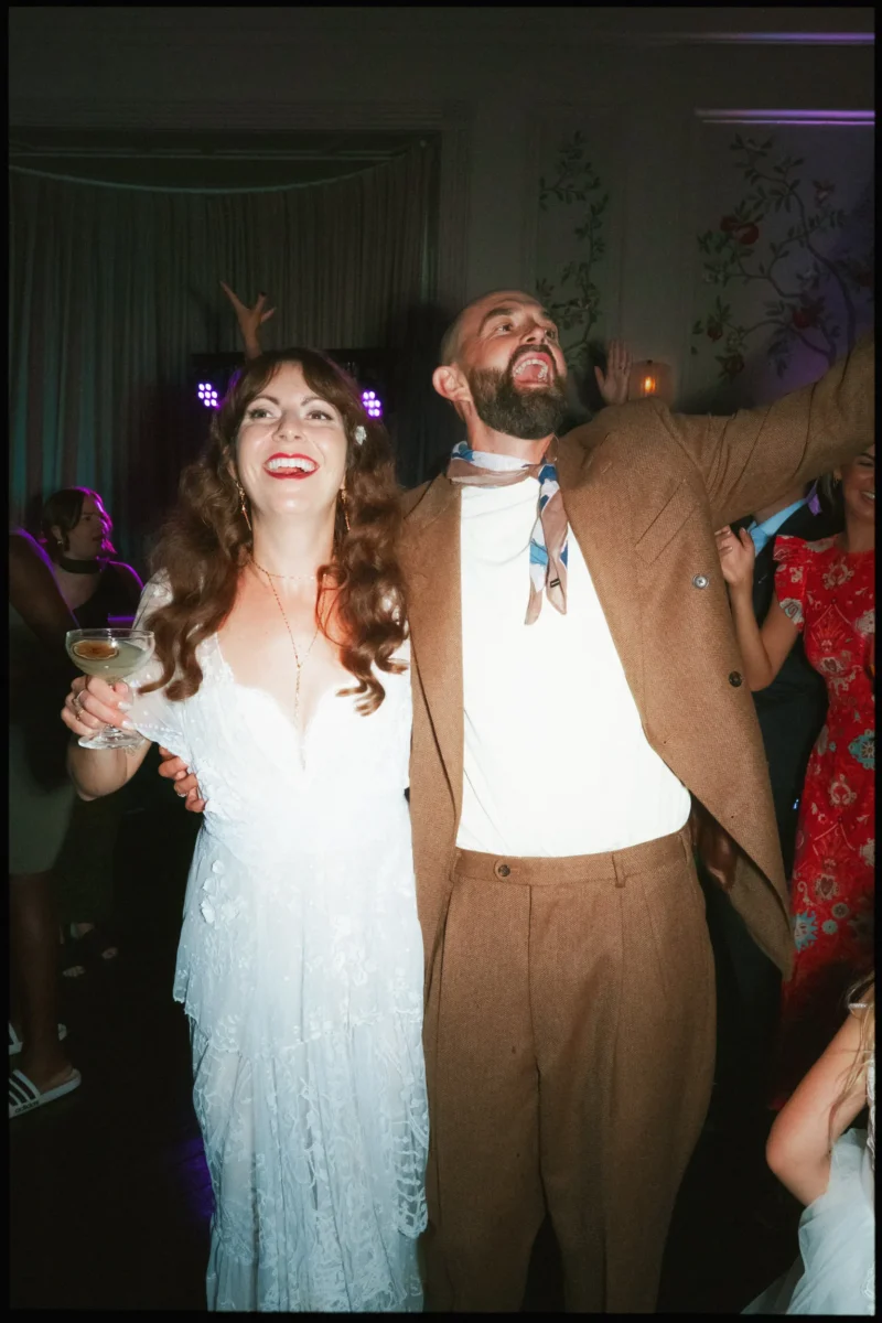 Bride in lace dress grins, coupe in hand, as her bearded groom throws an arm up on the dancefloor