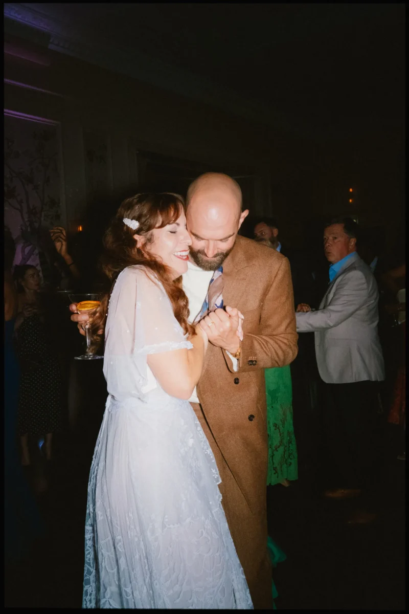 Smiling bride in a pale lace dress dances close with her bearded groom in a brown suit, cocktail in hand, at The George