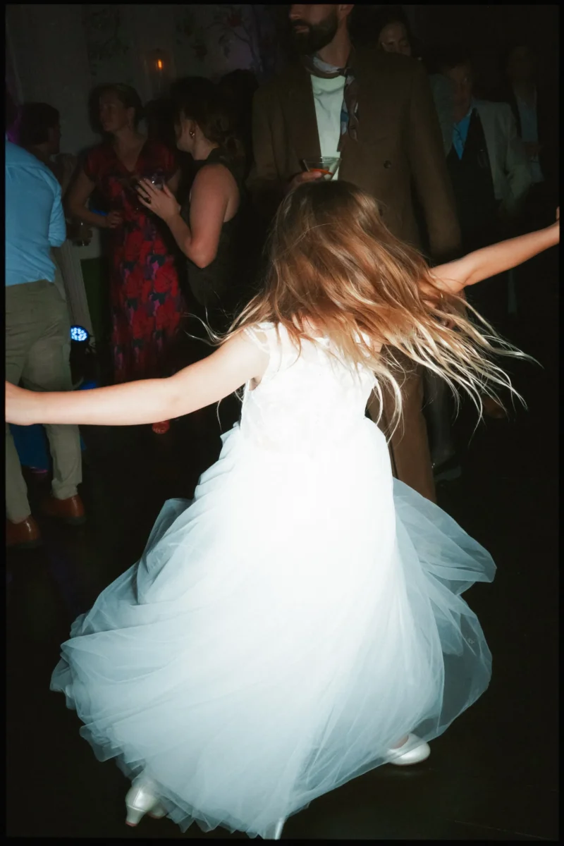 Flower girl in a white tulle dress spins with arms outstretched, hair flying, as guests chat on the dancefloor behind