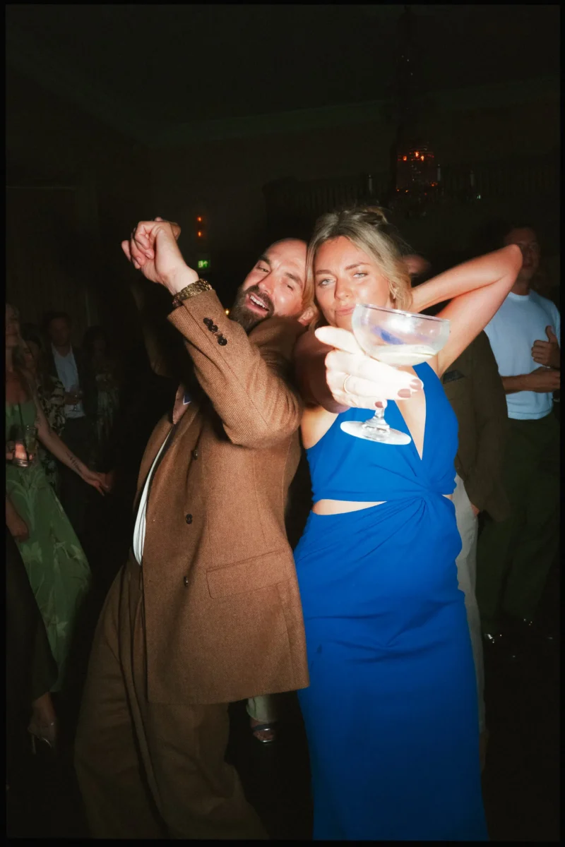 Guest in a blue dress thrusts out a coupe glass beside a laughing bearded man dancing in The George Ballroom