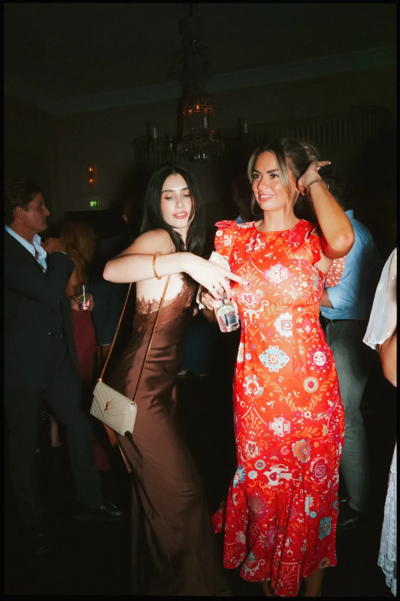 Two guests dance in a dimly lit room under a chandelier, one in a brown satin dress, the other in red florals