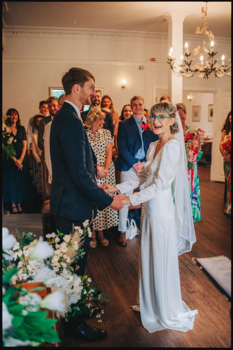 Bride in a silk gown and teal glasses laughs holding hands with her groom as guests beam behind them at the ceremony
